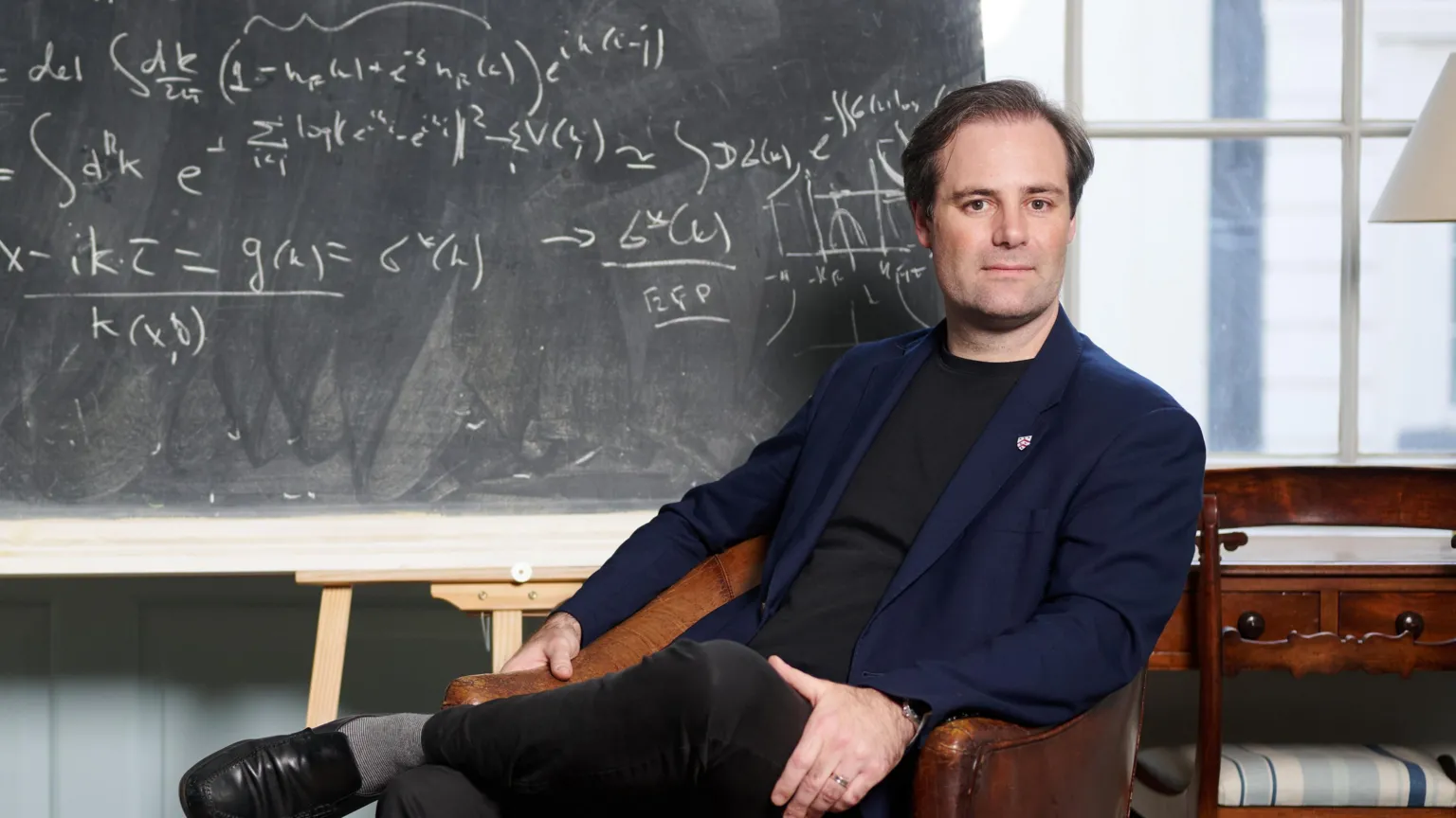 Ben Delo Foundation Ben Delo sits in front of a blackboard in a brown leather armchair. He has short brown hair and wears a blue blazer, black t-shirt, black trousers, grey socks and black loafers. A maths equation is written on the board behind him.