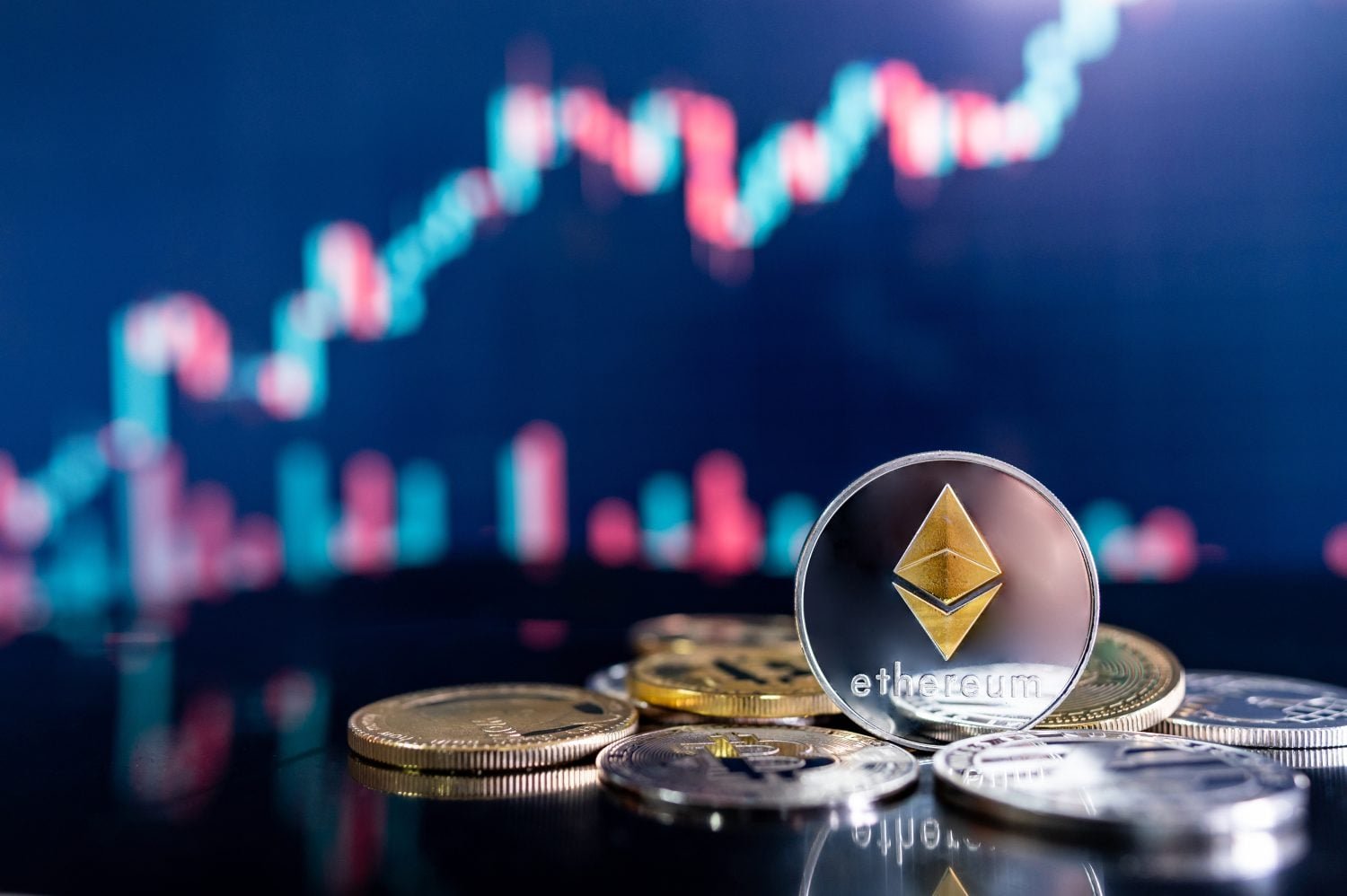 A silver Ethereum cryptocurrency coin with a gold logo and 'ethereum' text is placed prominently on a dark, reflective surface, surrounded by other gold and silver digital coins. In the background, a blurred electronic display shows a financial market chart with red and blue upward and downward trends.
