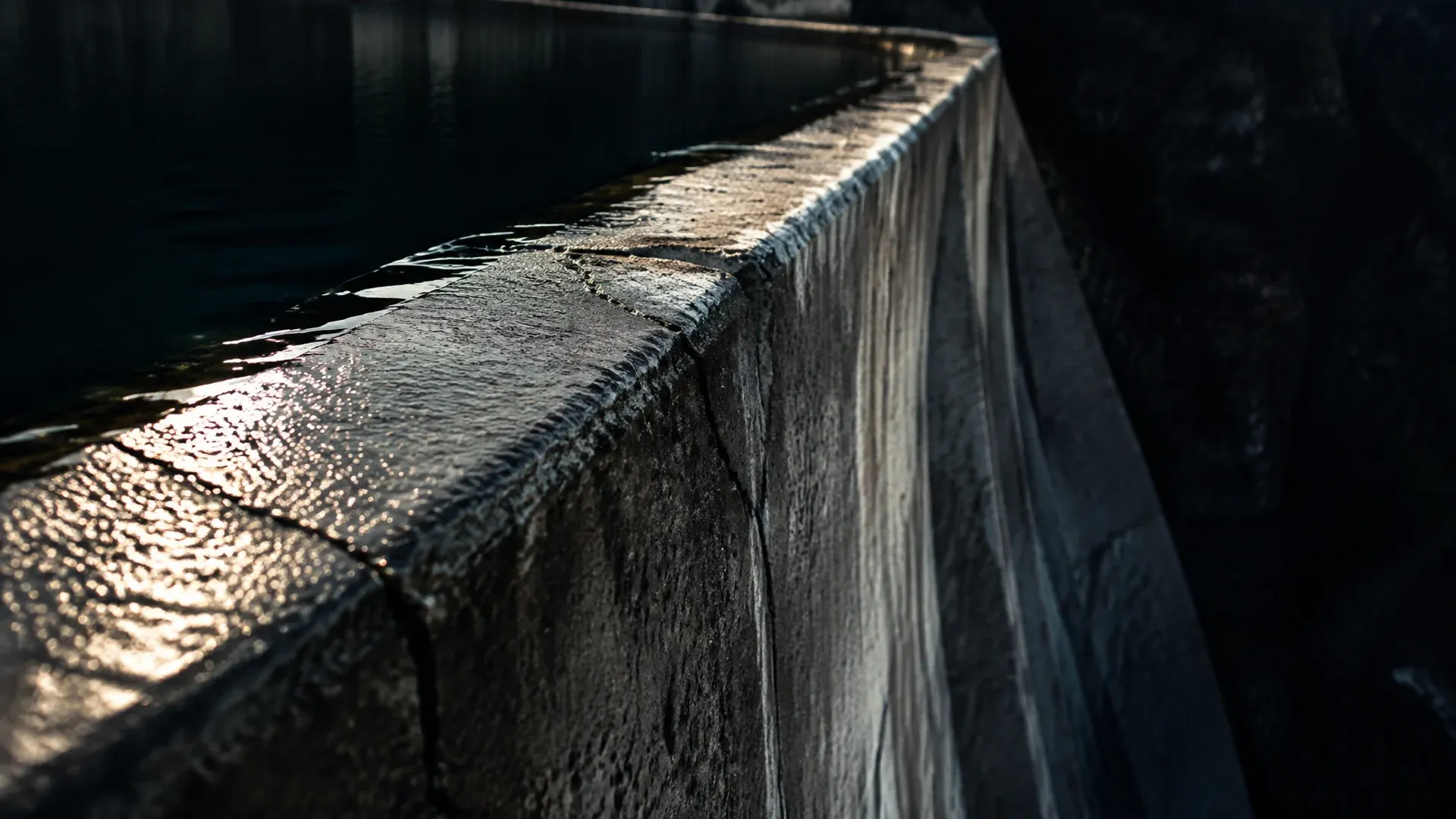Concrete crest of a weir in raking light, the dark water standing a few centimetres below the edge and lying still